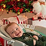 baby, smiling, infant, baby_seat, christmas_tree, christmas_lights, ornaments, santa_decoration, holiday, festive, gift_boxes, red, green, indoors, happy, child, cute, seasonal, cozy, celebration