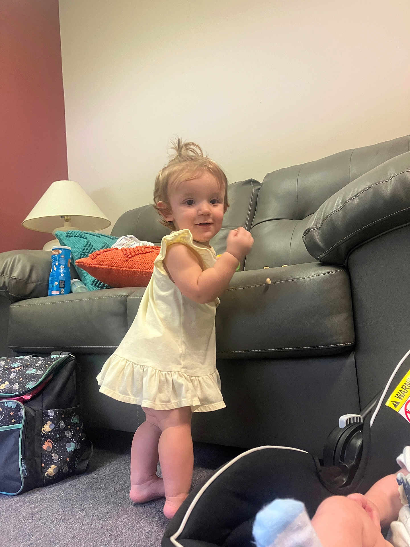 Ava is registered to the contest to win money with this photo: toddler, child, baby, standing, dress, indoor, carpet, couch, pillow, lamp, baby_car_seat, footwear, hair, person, room, toy, bag, floor, expression, furniture