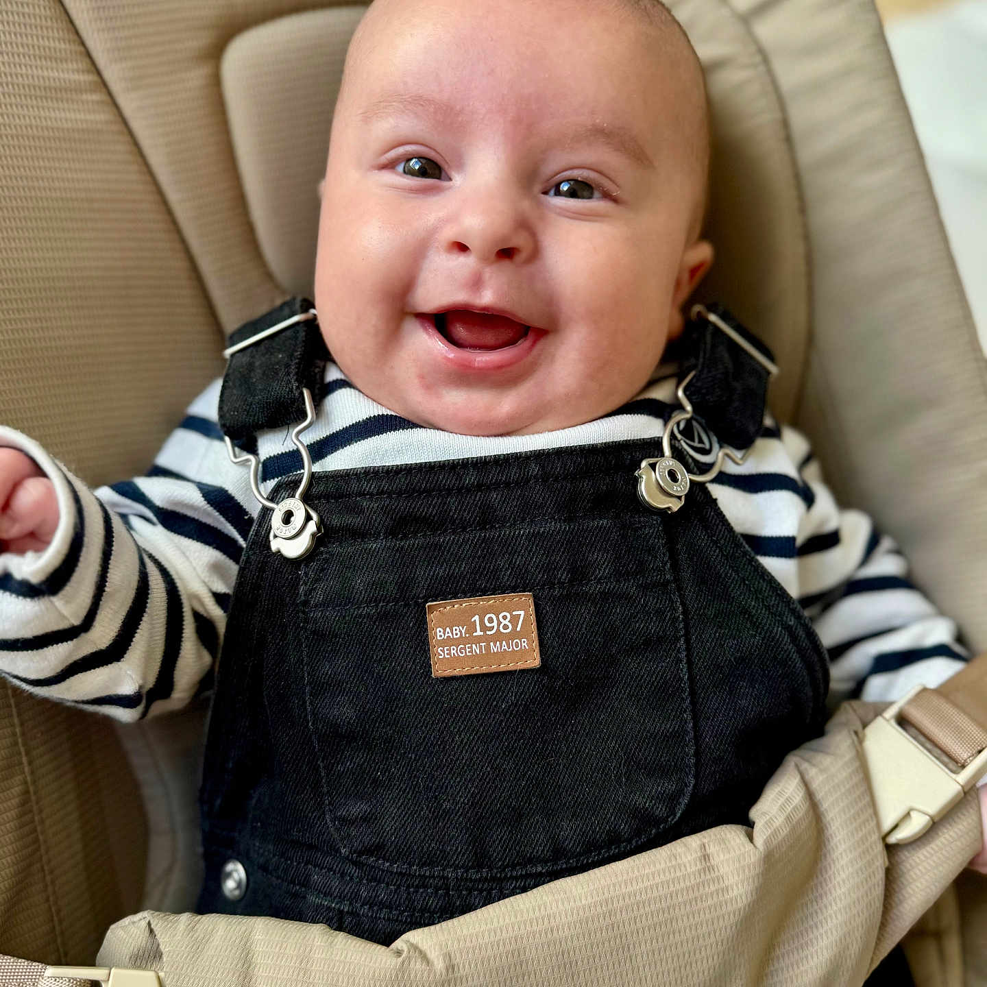 Arthur participe au concours pour gagner de l'argent avec cette photo : baby, baby_carrier, baby_fashion, child, closeup, clothing, cute, expression, face, happy, indoor, infant, overalls, person, portrait, seat, sitting, smiling, striped_shirt, young_child