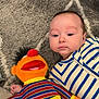 Noah is registered to the contest to win money with this photo: baby, child, plush_toy, doll, striped_clothing, blue, orange, red, yellow, face, person, carpet, floor, hand, toy, indoor, cute, soft, lying_down, playtime