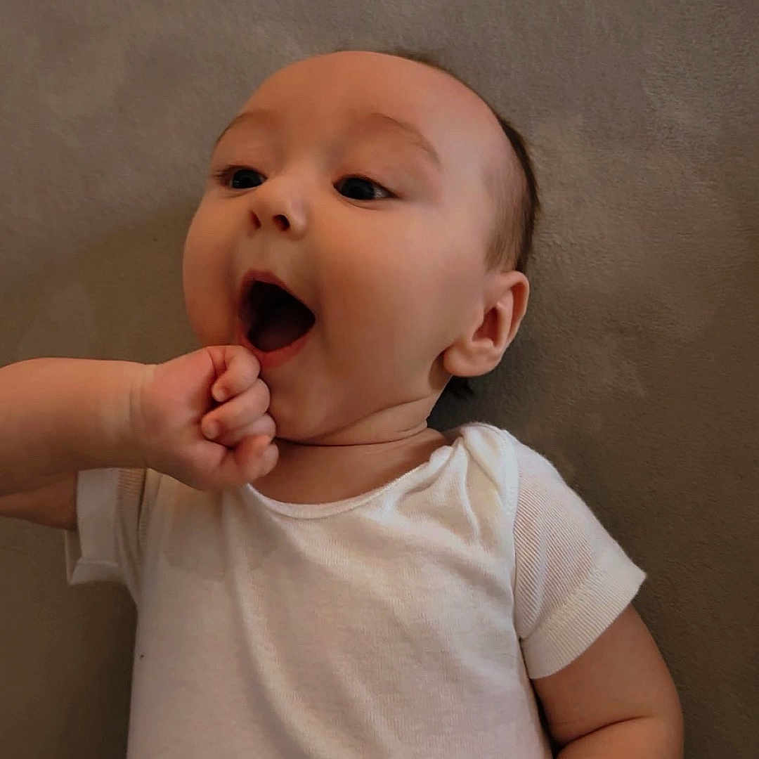 Noah joined the competition — help win amazing prizes! adorable, arm, baby, child, cute, expression, fist, hand, head, indoors, infant, lying_down, mouth_open, onesie, person, portrait, skin, soft_surface, white_clothing, young