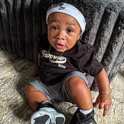 Caizen Knox is registered to the contest to win money with this photo: toddler, child, curly_hair, headband, nike, black_shirt, gray_shorts, sneakers, carpet, couch, indoor, jewelry, necklace, person, portrait, sitting, cute, expression, floor, furniture