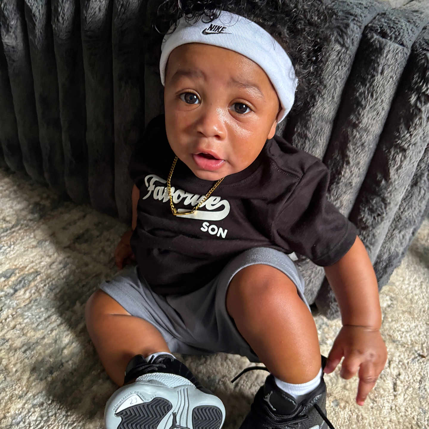 Caizen Knox is registered to the contest to win money with this photo: toddler, child, curly_hair, headband, nike, black_shirt, gray_shorts, sneakers, carpet, couch, indoor, jewelry, necklace, person, portrait, sitting, cute, expression, floor, furniture