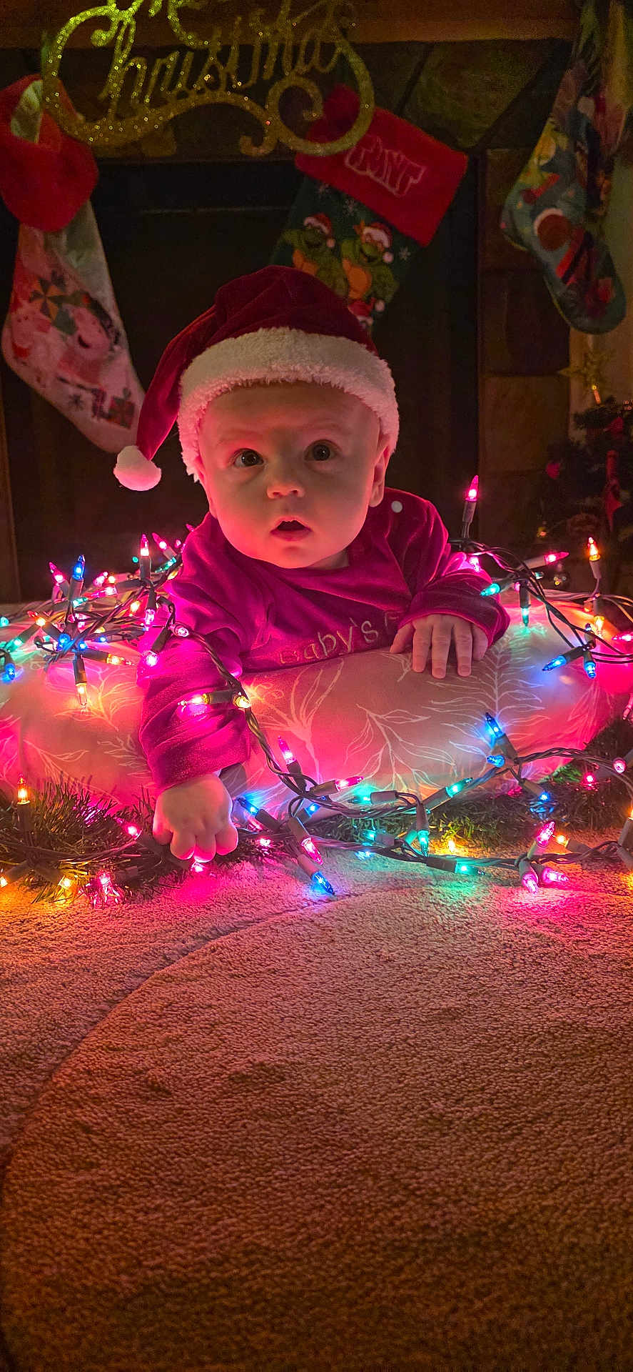 James is registered to the contest to win money with this photo: baby, child, christmas, holiday, festive, santa_hat, lights, colorful_lights, cushion, carpet, fireplace, stockings, indoor, cute, portrait, holiday_decor, celebration, festivities, toddler, curious
