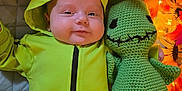 James joined the competition — help win amazing prizes! baby, child, costume, green_clothing, knitted_toy, plush_toy, smile, face, cute, autumn_leaves, lights, soft_toy, indoor, quilted_surface, holiday, festive, cozy, portrait, infant, cuddling
