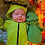 baby, child, costume, green_clothing, knitted_toy, plush_toy, smile, face, cute, autumn_leaves, lights, soft_toy, indoor, quilted_surface, holiday, festive, cozy, portrait, infant, cuddling
