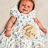 baby, infant, child, smiling, dress, butterfly_pattern, hair_bow, wooden_sign, milestone, lying_down, cute, soft_background, portrait, skin, hand, face, happy, young_child, white_dress, three_months