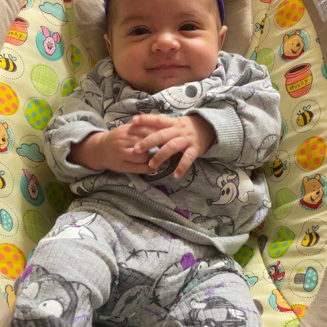 Heaven is registered to the contest to win money with this photo: baby, infant, child, smiling, portrait, sitting, bouncer, baby_seat, purple_bow, gray_outfit, cartoon_clothing, hands, indoors, cozy, colorful, winnie_the_pooh, cushion, close_up, happy, photography