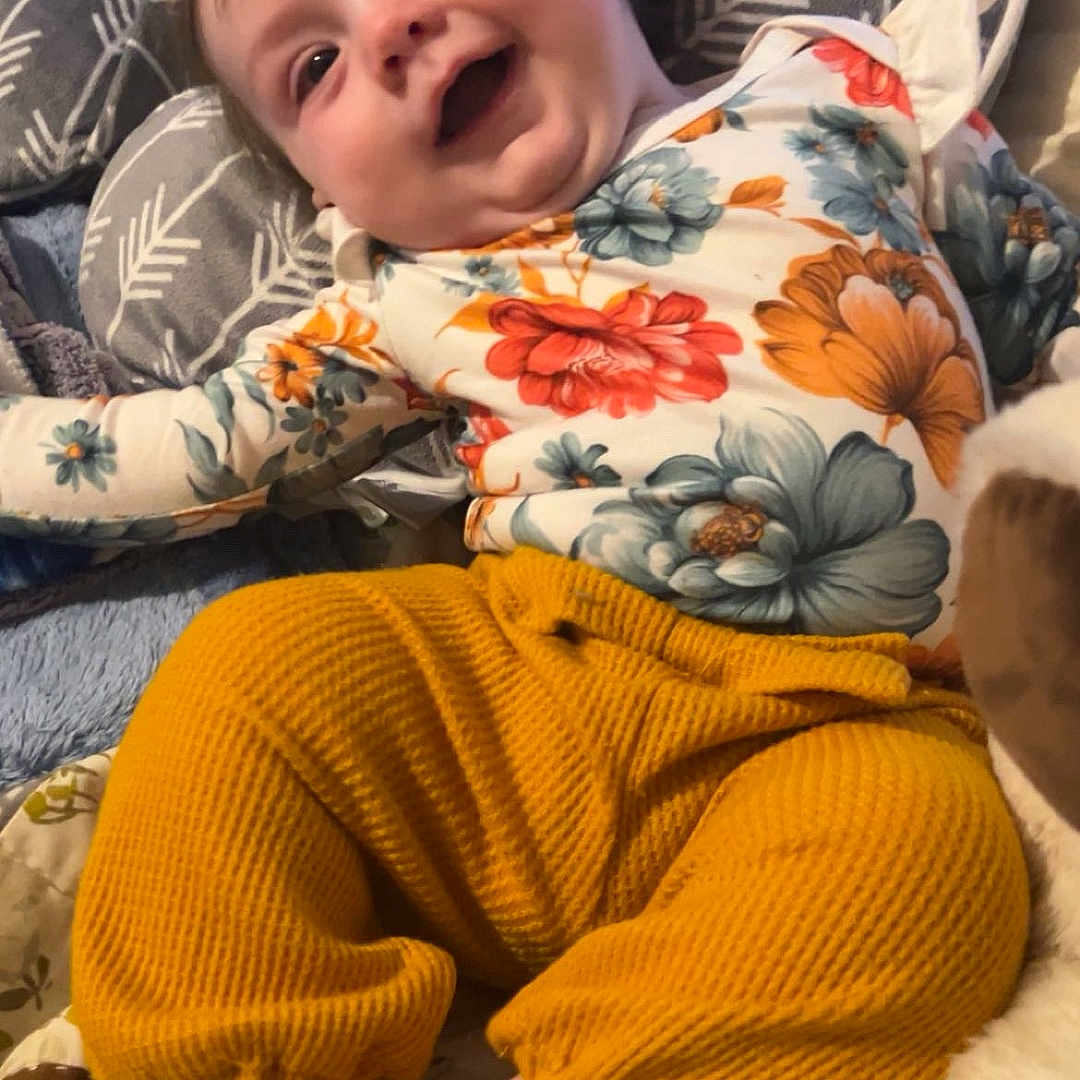 Ryleigh is registered to the contest to win money with this photo: baby, child, clothing, comfort, cushion, cute, floral_shirt, foot, happy, indoor, infant, pattern, playful, plush, portrait, relaxed, smiling, sock, soft_toy, yellow_pants