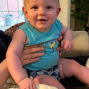 Levi is registered to the contest to win money with this photo: baby, child, infant, smiling, blue_shirt, checkered_shorts, blanket, hand, indoor, door, glass_door, plant, couch, person, face, happy, blonde_hair, seated, home_interior, toddler