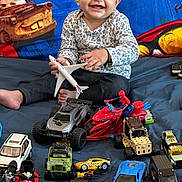 Navisha joined the competition — help win amazing prizes! baby, child, smiling, toy_airplane, toy_cars, bed, blanket, vehicles, indoor, playtime, happy, person, blue, red, yellow, car, motorcycle, spiderman_toy, clothing, floor