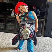 Ethan is registered to the contest to win money with this photo: child, boy, red_cowboy_hat, life_vest, skull_costume, peace_sign, barefoot, carpet, living_room, television, cabinet, toy, smiling, playful, indoor, furniture, person, blue_pants, reflection, photography