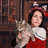 woman, cat, tabby_cat, holiday, christmas, festive, headband, reindeer, sweater, red, white, wooden_shelf, toy_truck, teddy_bear, window, lights, indoor, portrait, holding, decorations