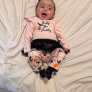 Layla is registered to the contest to win money with this photo: baby, infant, child, smiling, happy, bed, white_bedspread, pink_clothing, halloween_theme, pumpkin, ghost, bat, black_shoes, toddler, cute, lying_down, patterned_pants, headband, indoors, celebration