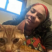 Moses Allen is registered to the contest to win money with this photo: person, cat, orange_tabby, indoor, window, curly_hair, headband, smile, holding, closeup, arm, casual_clothing, necklace, face, pet, domestic_animal, feline, light, background, portrait
