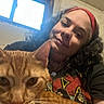 person, cat, orange_tabby, indoor, window, curly_hair, headband, smile, holding, closeup, arm, casual_clothing, necklace, face, pet, domestic_animal, feline, light, background, portrait