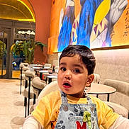 Ghali participe au concours pour gagner de l'argent avec cette photo : toddler, child, high_chair, restaurant, mural, art, colorful, denim, overall, mickey_mouse, pacifier, table, chair, indoor, casual_clothing, curly_hair, baby, face, expression, interior