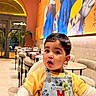 toddler, child, high_chair, restaurant, mural, art, colorful, denim, overall, mickey_mouse, pacifier, table, chair, indoor, casual_clothing, curly_hair, baby, face, expression, interior