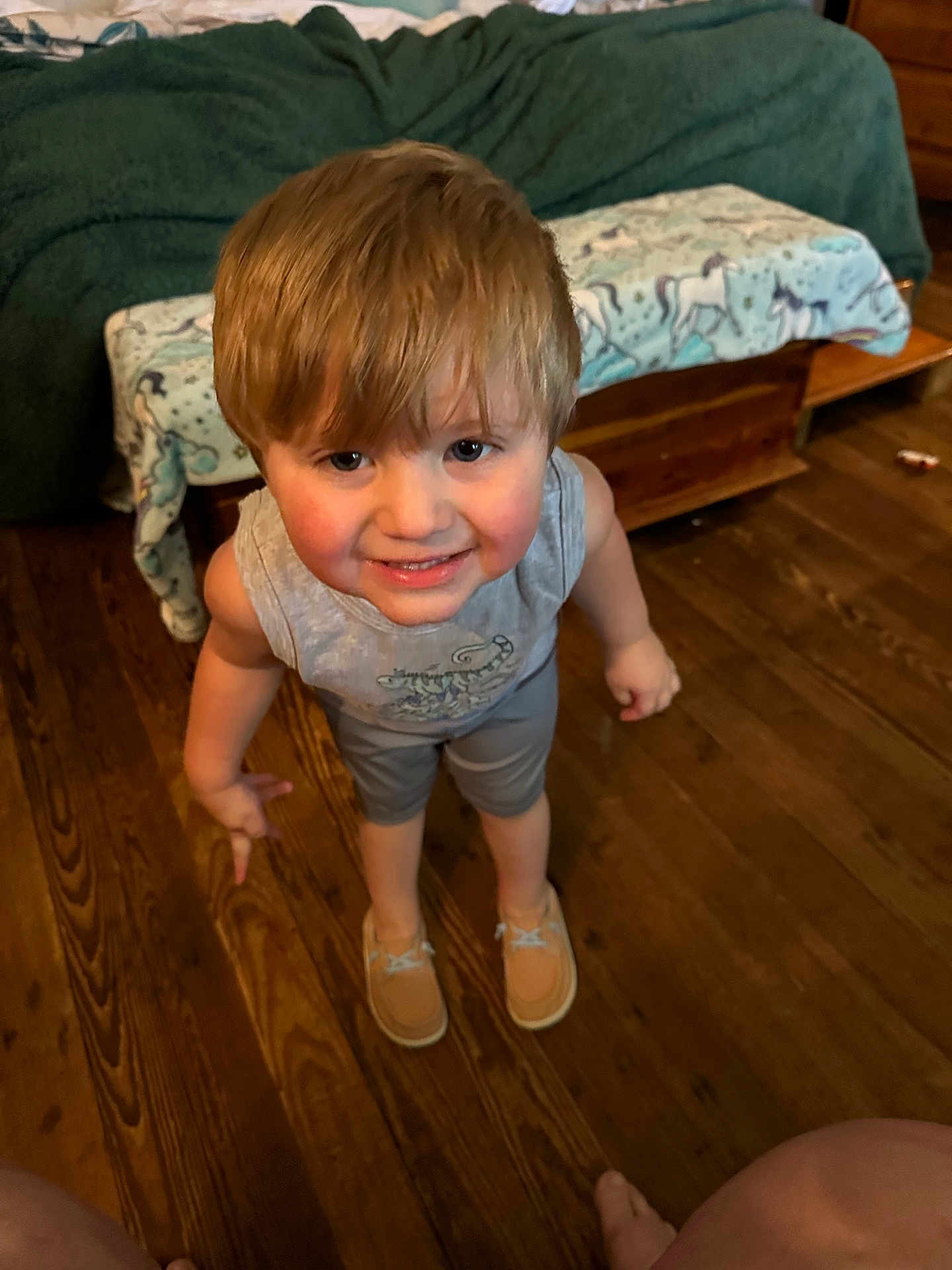 Karson is registered to the contest to win money with this photo: child, smiling, blonde_hair, indoor, wooden_floor, tank_top, shorts, shoes, person, looking_up, happy, home, blanket, furniture, bed, floor, casual_clothing, cute, young, portrait