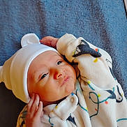 Ossie Schneider joined the competition — help win amazing prizes! baby, infant, child, white_hat, onesie, cozy, blanket, blue_background, cute, sleepwear, newborn, soft_fabric, patterned_clothing, portrait, resting, indoors, small_hands, face, expression, headwear