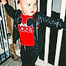 toddler, child, boy, mickey_mouse, red_shirt, black_jacket, sneakers, railing, concrete_floor, outdoor, night, curious, standing, casual_clothing, person, portrait, young_child, cute, fashion, expression