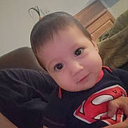 Ellie is registered to the contest to win money with this photo: baby, child, superman_shirt, indoor, person, cute, smiling, face, hand, hair, curly_hair, toddler, closeup, portrait, couch, blanket, cozy, skin, young_child, happy