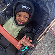 Kamari is registered to the contest to win money with this photo: toddler, child, stroller, hat, clothing, baby, hand, smile, face, urban, outdoor, jacket, hood, fabric, seatbelt, wheel, pavement, person, baby_bib, black_clothing
