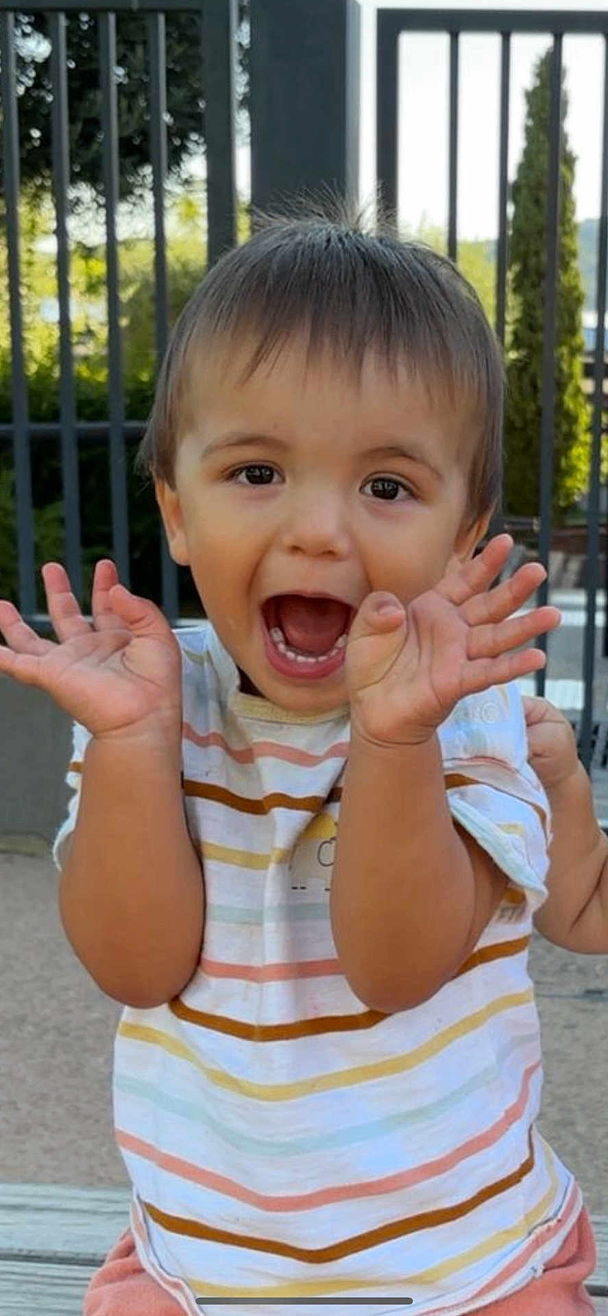 Eden participe au concours pour gagner de l'argent avec cette photo : bench, casual_clothing, child, daylight, expression, face, fence, greenery, hair, hands, happy, nature, outdoor, person, playful, shorts, smiling, striped_shirt, summer, toddler