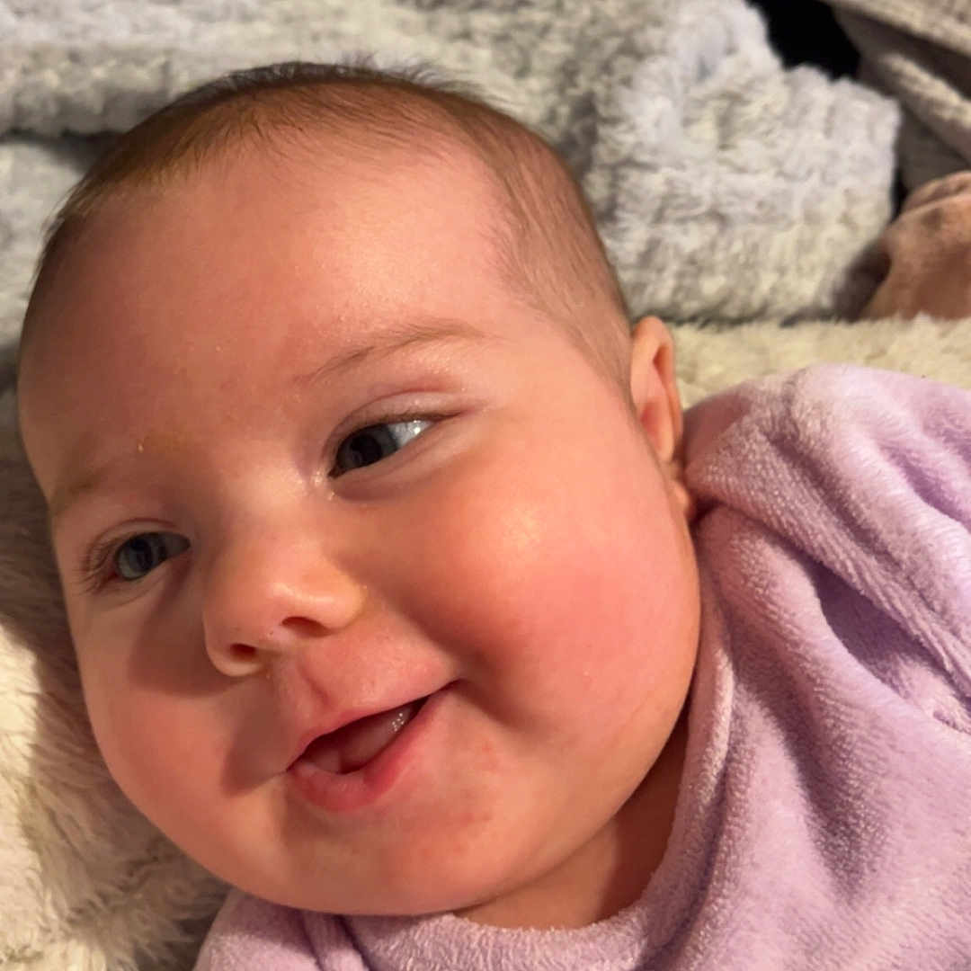 Elyana participe au concours pour gagner de l'argent avec cette photo : baby, blanket, cheeks, closeup, cozy, cute, expression, face, happy, head, indoors, infant, love_text, person, purple_clothing, skin, smiling, soft_texture, toddler, warm