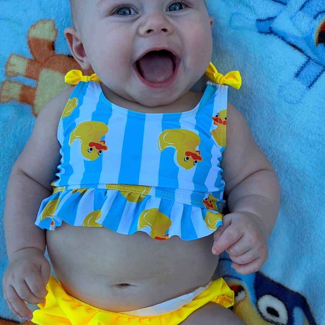 Delylah is registered to the contest to win money with this photo: baby, child, smiling, happy, blue, yellow, clothing, blanket, cartoon, pattern, face, infant, cute, skin, person, outdoor, lying_down, cheerful, expression, playful