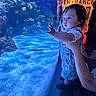 child, toddler, aquarium, glass, underwater, fish, blue_light, curiosity, hands, support, indoor, person, baby, overalls, reflection, water, marine_life, exhibit, face, expression