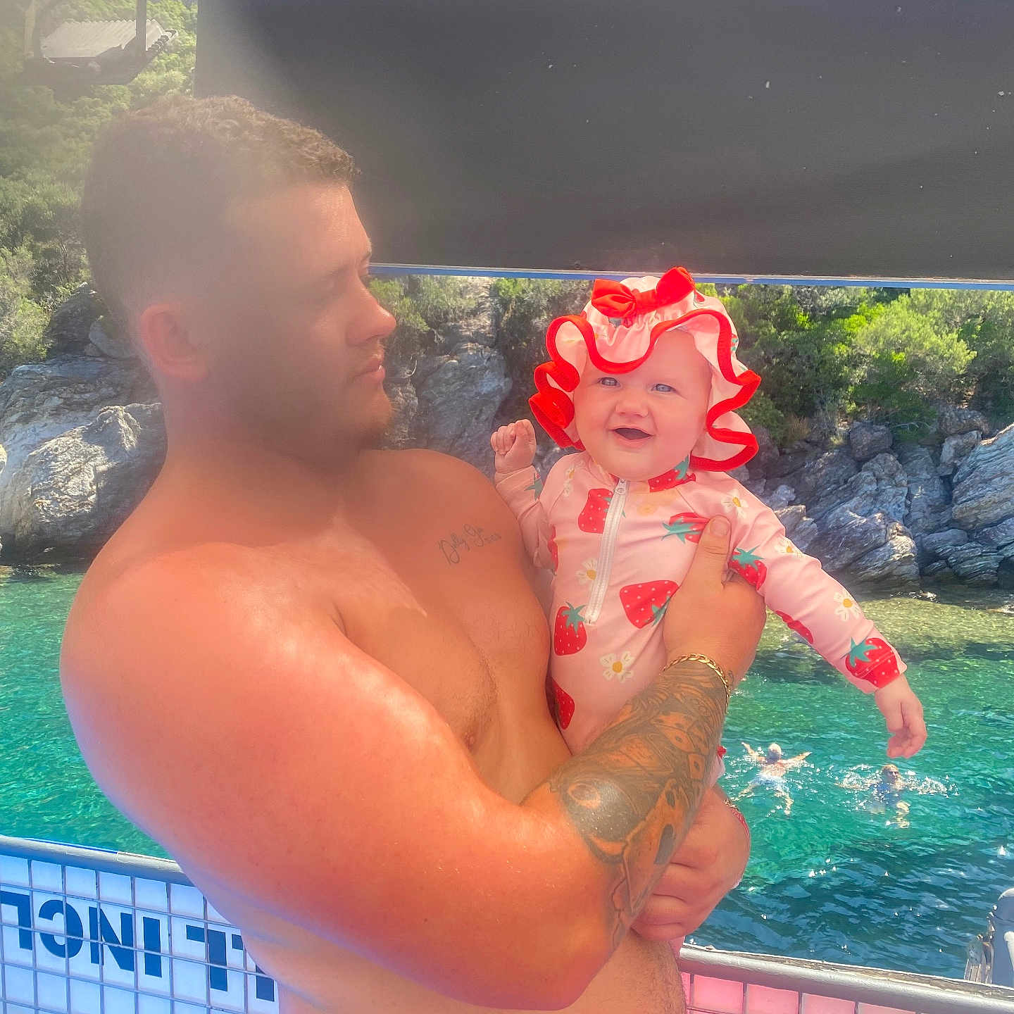 Dolly-Gee is registered to the contest to win money with this photo: baby, boat, child, happy, hat, holding, man, nature, outdoor, people, rocks, smile, summer, sunlight, swimming, swimsuit, tattoo, trees, vacation, water