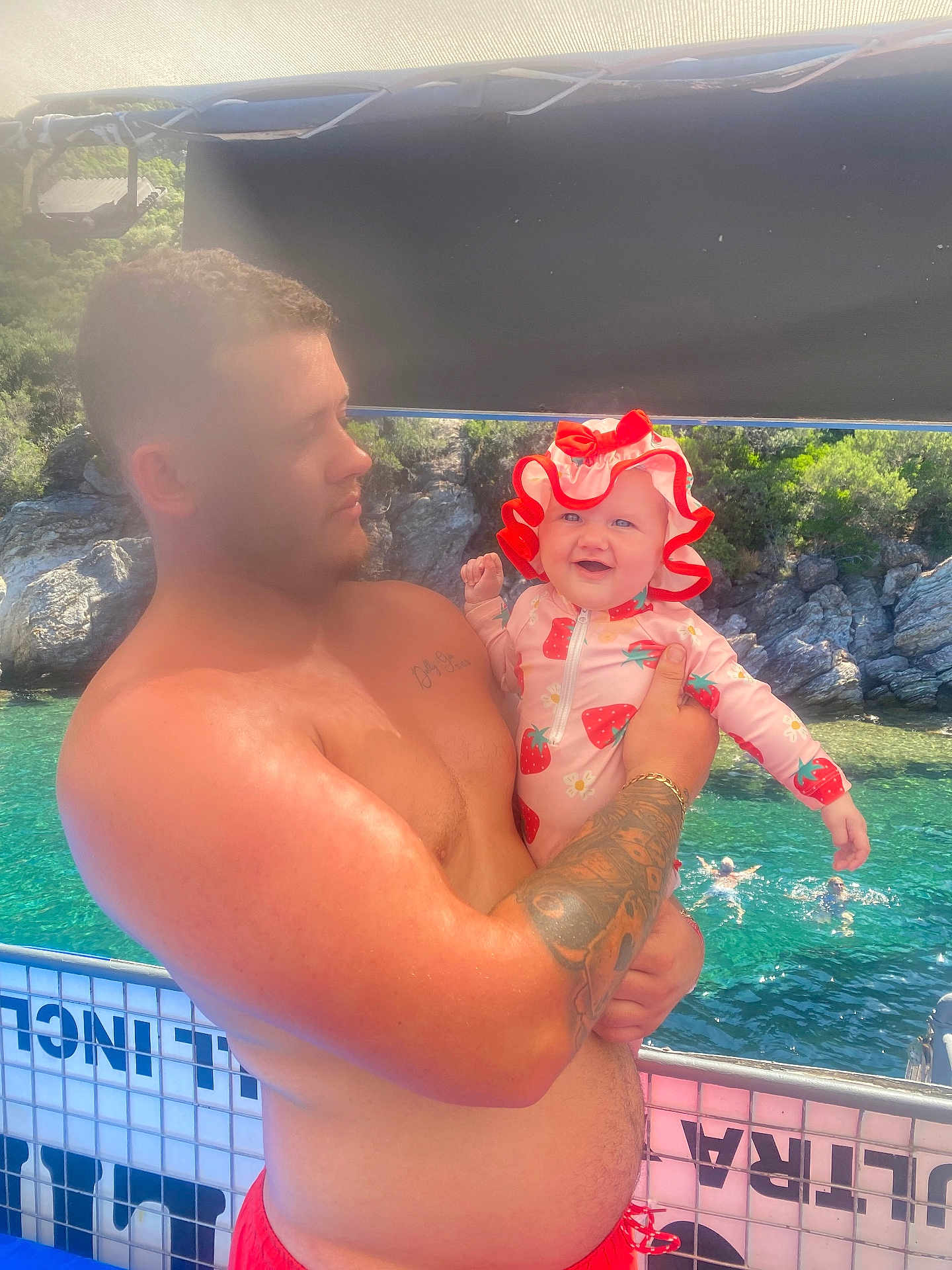 Dolly-Gee is registered to the contest to win money with this photo: man, baby, tattoo, swimsuit, hat, boat, water, swimming, rocks, trees, outdoor, sunlight, holding, child, smile, nature, summer, vacation, people, happy