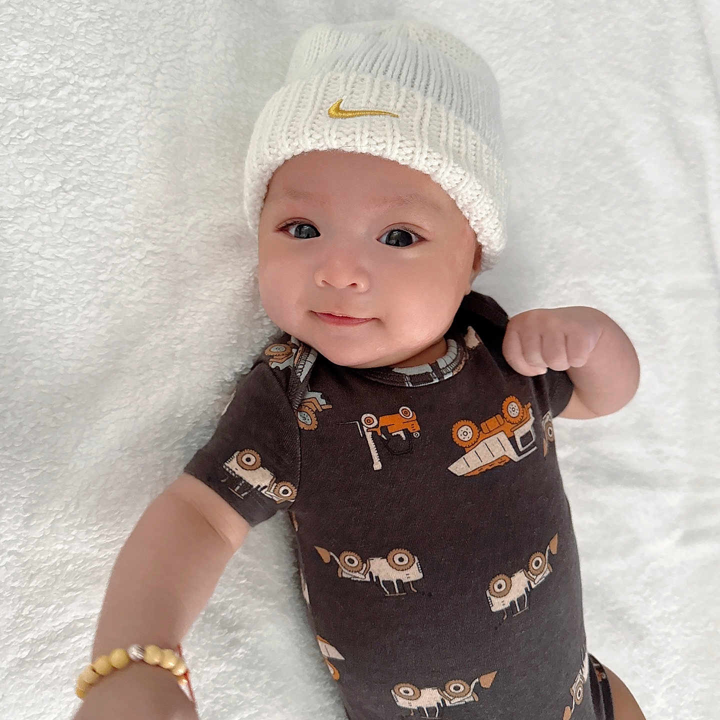 Barron is registered to the contest to win money with this photo: adorable, apparel, baby, blanket, bracelet, child, cozy, cute, face, hat, indoors, infant, knit_hat, lying_down, onesie, patterned_clothing, portrait, smiling, soft_texture, white_blanket
