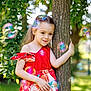 child, girl, tree, dress, red_dress, floral_pattern, bubbles, outdoor, greenery, nature, smiling, happy, sunlight, garden, portrait, long_hair, playful, summer, colorful, cute
