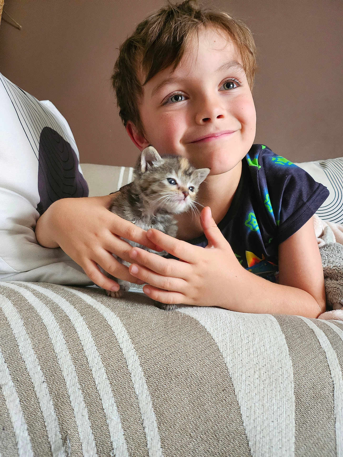 Cheshire participe au concours pour gagner de l'argent avec cette photo : child, kitten, cat, couch, striped_blanket, smiling, hands, indoor, cozy, young_boy, cute, pet, furniture, portrait, happy, casual_clothing, curly_hair, home, resting, animal