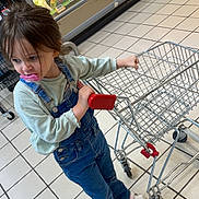 Chelsea participe au concours pour gagner de l'argent avec cette photo : casual_clothing, child, curious, denim, floor_tiles, footwear, grocery_store, indoor, long_sleeve_shirt, overalls, pacifier, person, produce, refrigerator_case, shopping, shopping_cart, supermarket, toddler, toy_cart, young_child