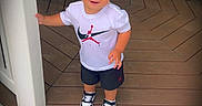 Lenox is registered to the contest to win money with this photo: toddler, child, smiling, portrait, standing, outdoor, porch, deck, doorway, doorframe, chairs, table, wooden_floor, sneakers, shorts, t_shirt, jordan_logo, blond_hair, sunlight, home