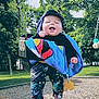 baby, child, swing, playground, park, trees, grass, smile, drool, bucket_swing, swing_chains, hat, blanket, colorful_clothing, patterned_pants, bare_feet, wood_chips, toy_key, sunny, outdoor