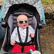 Cole is registered to the contest to win money with this photo: baby, infant, stroller, sunglasses, smile, seatbelt, canopy, outdoors, grass, stone_wall, cup_holder, sippy_cup, red_pants, jacket, portrait, happy, cute, toddler, baby_accessory, playful