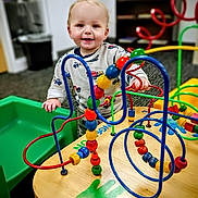 Cole is registered to the contest to win money with this photo: child, toddler, baby, smiling, bead_maze, wooden_table, playroom, toy, colorful, indoor, portrait, person, green_tray, car_pattern, hand, eyes, blond_hair, seat, carpet, beads