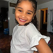 Delilah is registered to the contest to win money with this photo: casual_clothing, child, curly_hair, cute, earrings, girl, hand_on_hip, happy, home, indoor, light_skin, living_room, natural_light, person, portrait, posing, smiling, white_shirt, wooden_floor, young