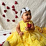 baby, blanket, celebration, child, cute, decor, fabric_flowers, golden_shoes, happy, headband, heart_shape, indoors, portrait, red_rose, rose_petals, sitting, smiling, white_bow, yellow_dress, young_child