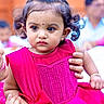 Aira is registered to the contest to win money with this photo: child, toddler, girl, portrait, pink_dress, traditional_clothing, hair_bun, curly_hair, bindi, serious_expression, brown_eyes, sitting, adult_hands, outdoor, crowd, blurred_background, colorful, fashion, jewelry, cultural_event