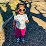 child, toddler, girl, sunglasses, outdoor, portrait, standing, smiling, fashion, headband, floral_shirt, pink_pants, sneakers, playground, path, gravel, park, sunlight, shadows, trees