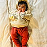Aira is registered to the contest to win money with this photo: baby, child, infant, portrait, smiling, lying_down, bed, bedding, hoodie, mittens, red_pants, knitwear, clothing, indoor, cozy, warm_lighting, happy, face, dark_hair, photography