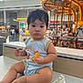 Kylo is registered to the contest to win money with this photo: baby, child, infant, snack, countertop, carousel, indoor, lights, food, clothing, person, face, seat, shopping_mall, blurred_background, curly_hair, cute, sitting, expression, toy