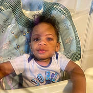 Giovanni is registered to the contest to win money with this photo: toddler, child, high_chair, face, person, curly_hair, indoor, clothing, shirt, expression, looking, infant, baby, young_child, seat, plastic_barrier, curiosity, home, light, portrait