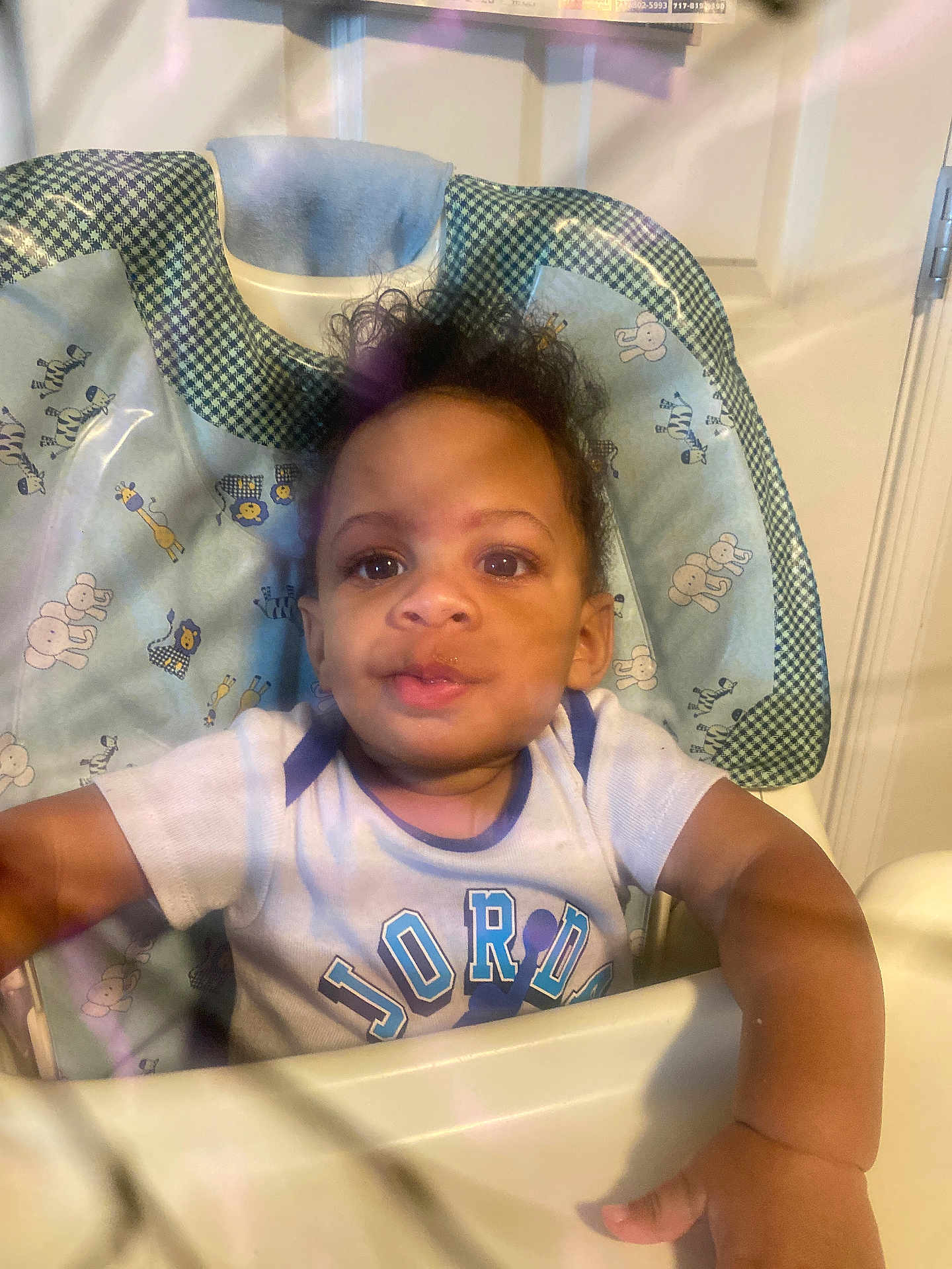 Giovanni is registered to the contest to win money with this photo: toddler, child, high_chair, face, person, curly_hair, indoor, clothing, shirt, expression, looking, infant, baby, young_child, seat, plastic_barrier, curiosity, home, light, portrait