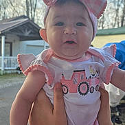 Isabelle is registered to the contest to win money with this photo: baby, child, pink_bow, headband, outdoor, person, cute, clothing, summer_wear, smiling, holding_hands, house, trees, expression, infant, portrait, daylight, face, cheeks, arms